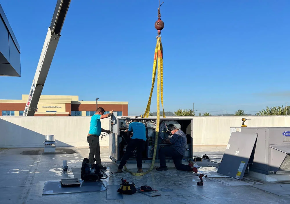 Three technicians work on a large HVAC unit being lifted by a crane on a commercial rooftop.